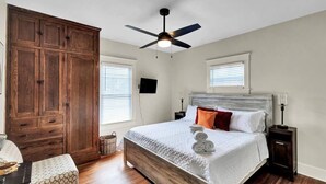 Ein Schlafzimmer mit großem Holzschrank, einem Bett mit weißer Bettwäsche, Deckenventilator und einem an der Wand montierten Fernseher.