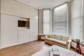 Ein modernes Schlafzimmer mit Holzboden, einem beigefarbenen Sofa, einem an der Wand montierten Fernseher und großen Fenstern mit Jalousien.
