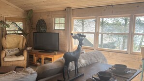 A cozy living room with a wooden ceiling, a television, a giraffe statue, and a dining area with a table set for two. - Cooperstown Lake Cabin Million Dollar Views (Cooperstown)