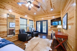 L’intérieur d’une cabane en rondins comprend un lit, deux fauteuils en cuir noir, une petite table, un téléviseur et un ventilateur de plafond.