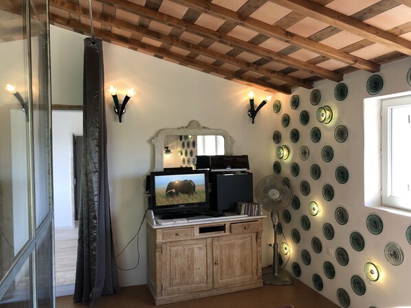 Quarto com teto de madeira, televisão sobre um móvel, ventoinha fixada na parede e pratos decorativos.