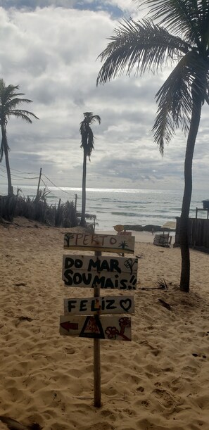 Un letrero de playa con mensajes en portugués, palmeras y el océano al fondo.