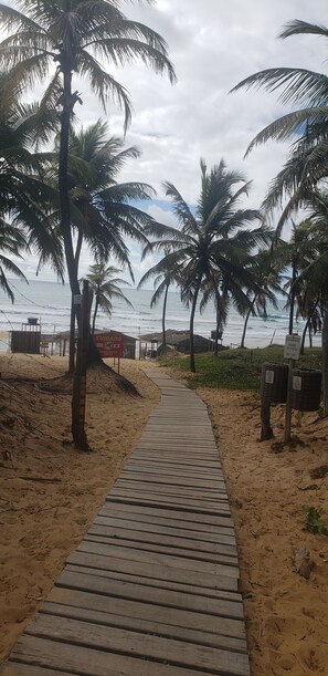 A wooden pathway leading to a beach with palm trees and a sign indicating a lifeguard station. - Estrela Antônia - Praia de Santo Antônio (Mata de São João)