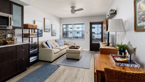 A modern apartment with a kitchenette, a living area with a sofa and ottoman, a dining table with chairs, and a large window with a view of buildings. - Sunset Beach Condo (Treasure Island)