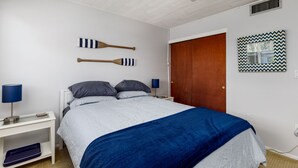 A bedroom with a bed, two wall-mounted paddleboards, a wooden door, a patterned wall decoration, and a nightstand with a lamp. - Sunset Beach Condo (Treasure Island)