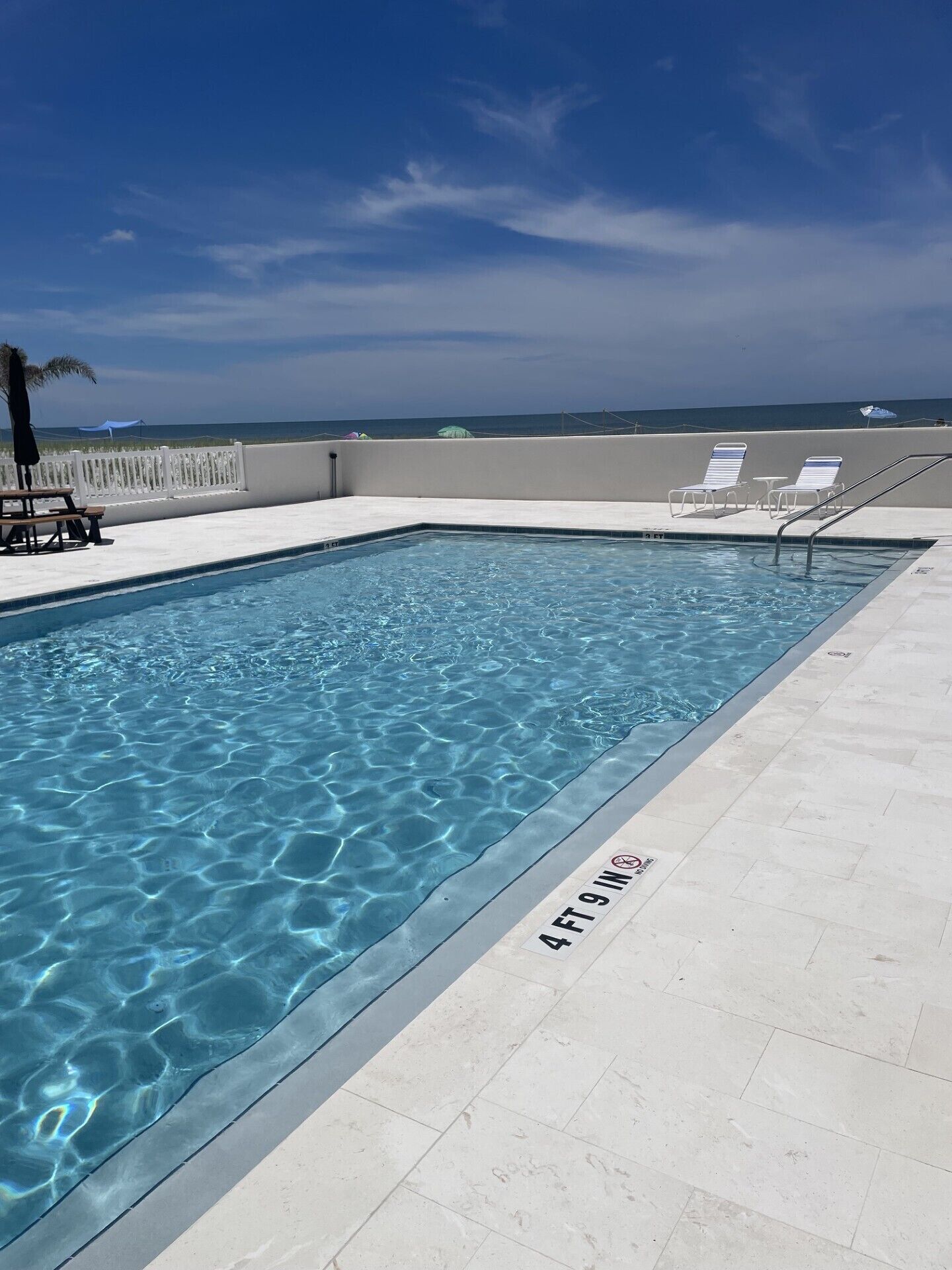 Ein rechteckiger Pool mit klarem, blauem Wasser, umgeben von einer weißen Liegefläche mit Liegestühlen und einem weißen Zaun.