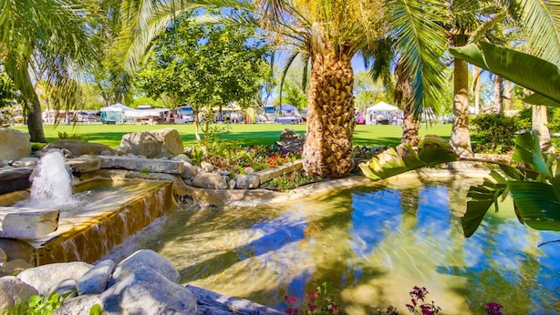 A tropical garden with a water feature, palm trees, and a grassy area with tents. - The Villas at Emerald Desert RV Resort (Palm Desert)