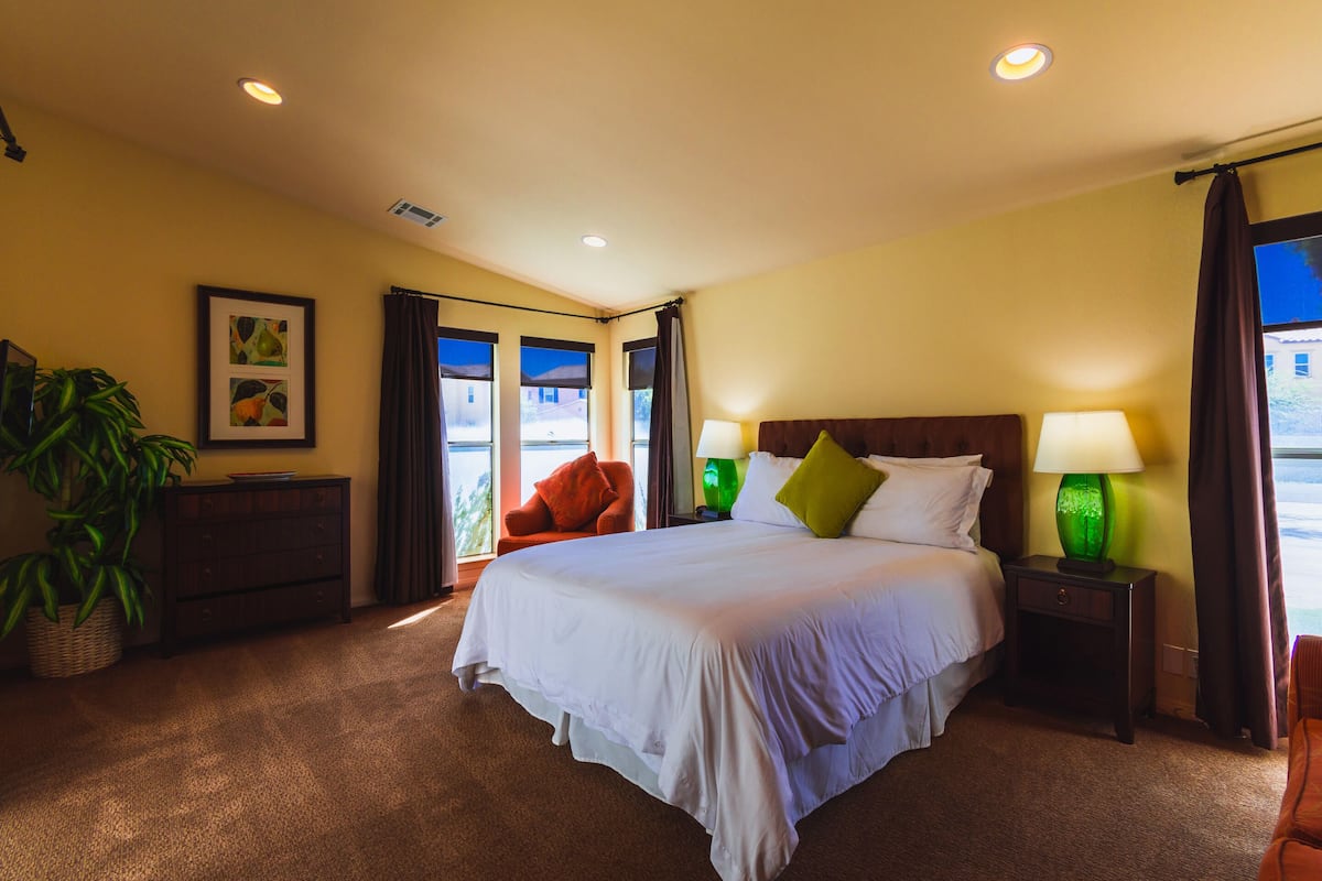 A hotel room with a large bed, two bedside lamps, a nightstand, a chair, a potted plant, and a window with curtains.