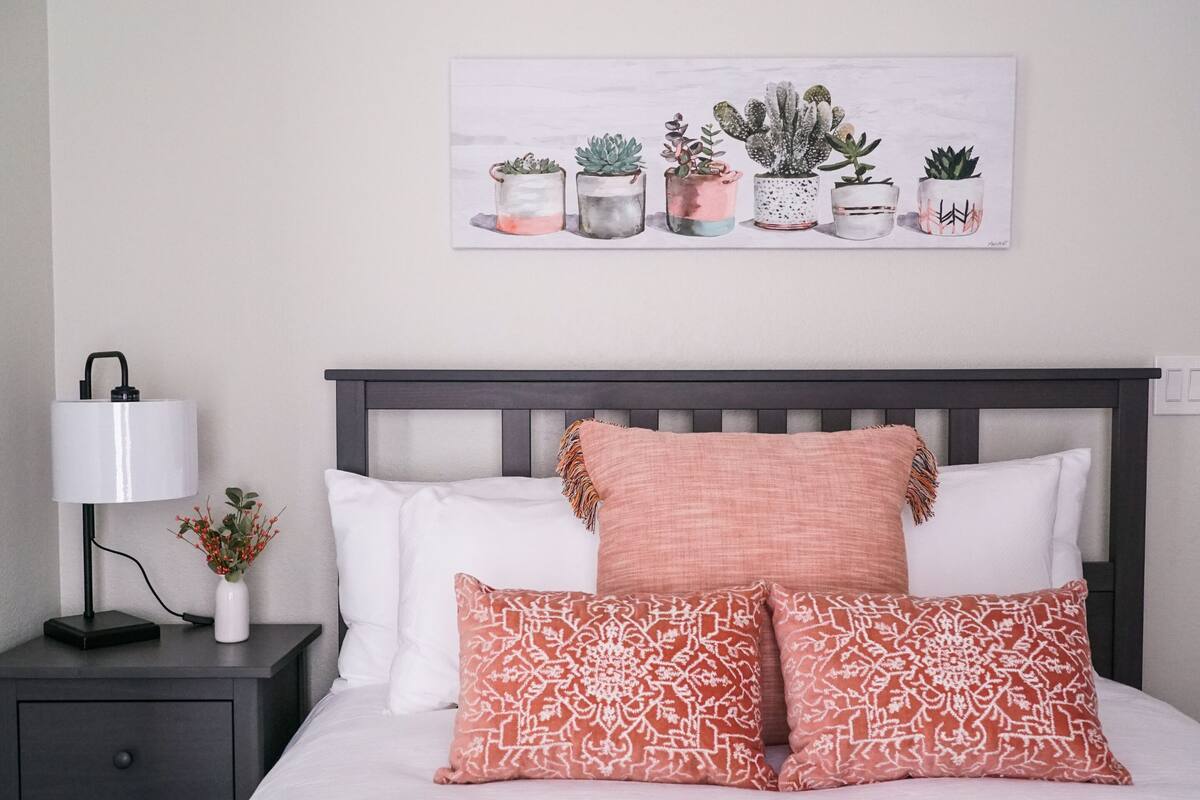 A neatly made bed with white and patterned pillows, a dark headboard, and a nightstand with a lamp and a vase of flowers.