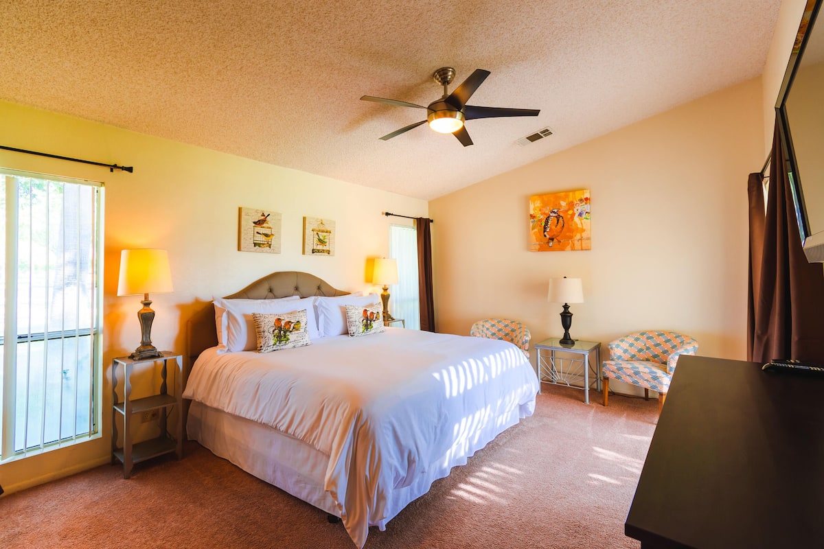 A hotel room with a large bed, two bedside tables with lamps, a ceiling fan, a window with curtains, and a painting on the wall.