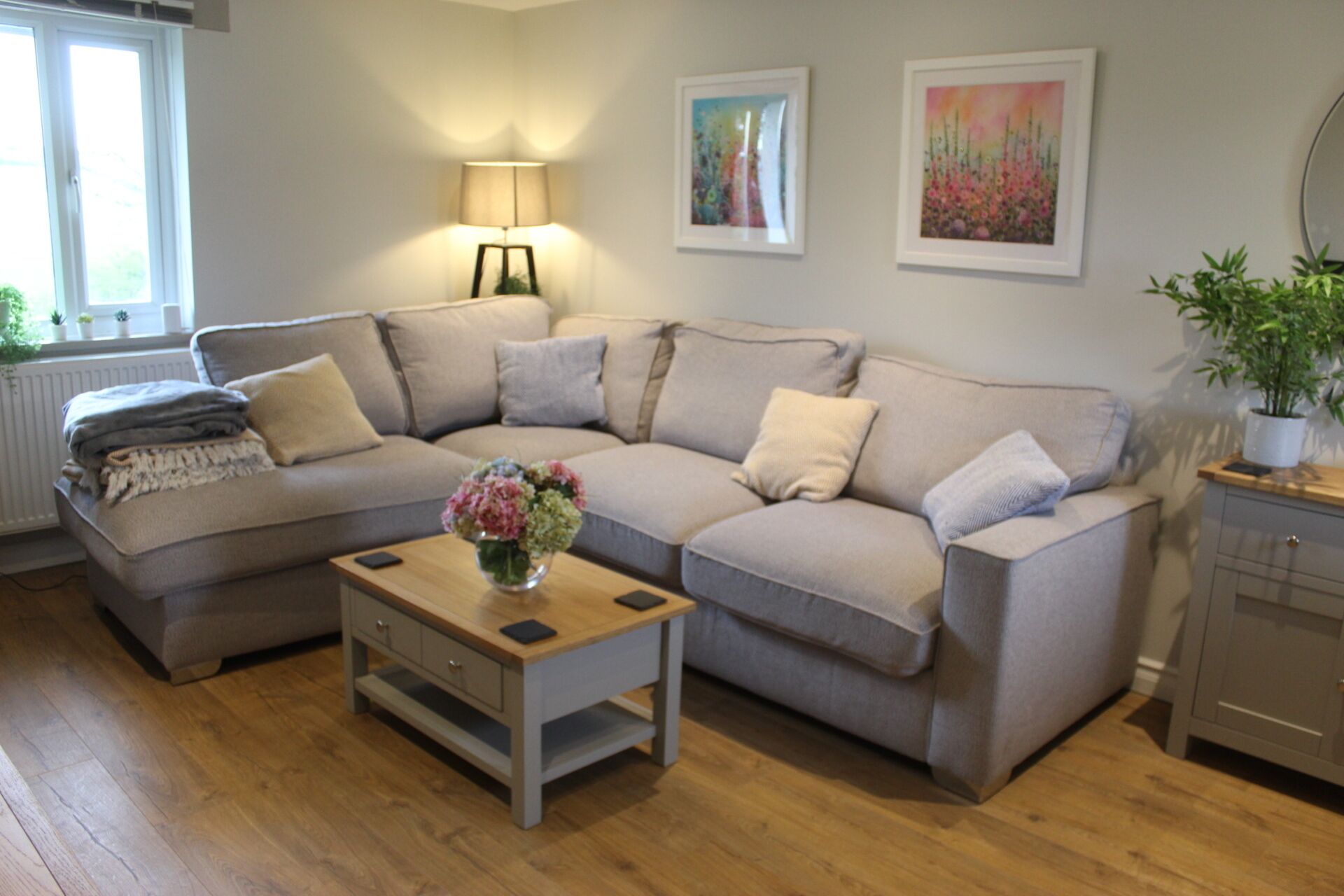 A modern living room with a sectional sofa, a coffee table, and a lamp. There are two framed artworks on the wall and a potted plant on a cabinet.