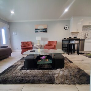 A modern living room with a glass coffee table, two red armchairs, a beige sofa, and a kitchen area with a hexagonal backsplash and stainless steel appliances. - Modern Residence in Torrance 15 mins from LAX (Torrance)