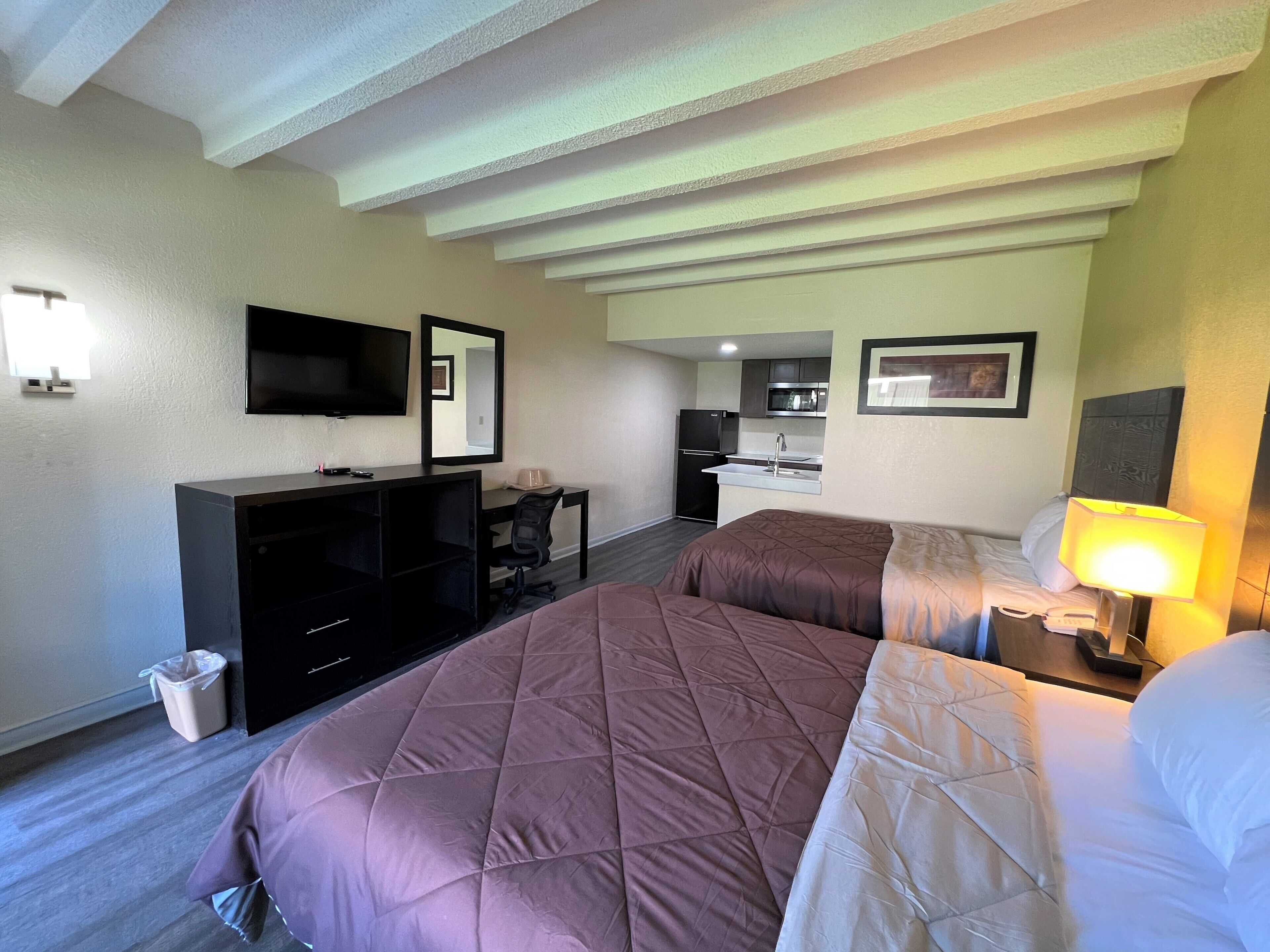A hotel room with two beds, a desk, a television, a mirror, a kitchen area, and a trash bin.