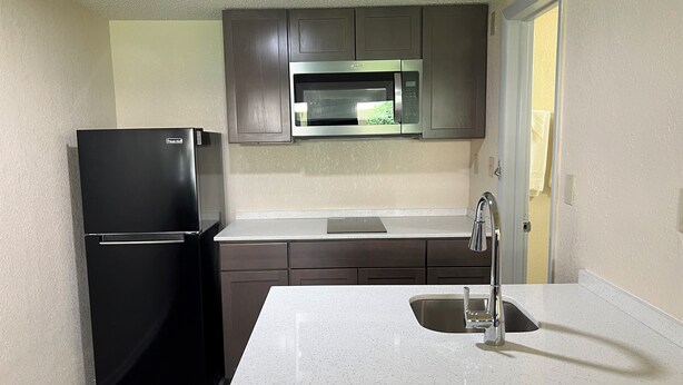 A compact kitchen with a black refrigerator, stainless steel microwave, and a white countertop with a sink. - James River Inn & Suites (Newport News)