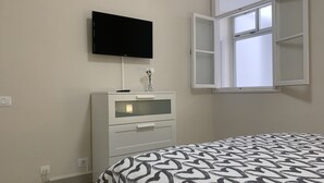 Een slaapkamer met een bed, een aan de muur bevestigde televisie, een dressoir en een raam met geopende luiken.