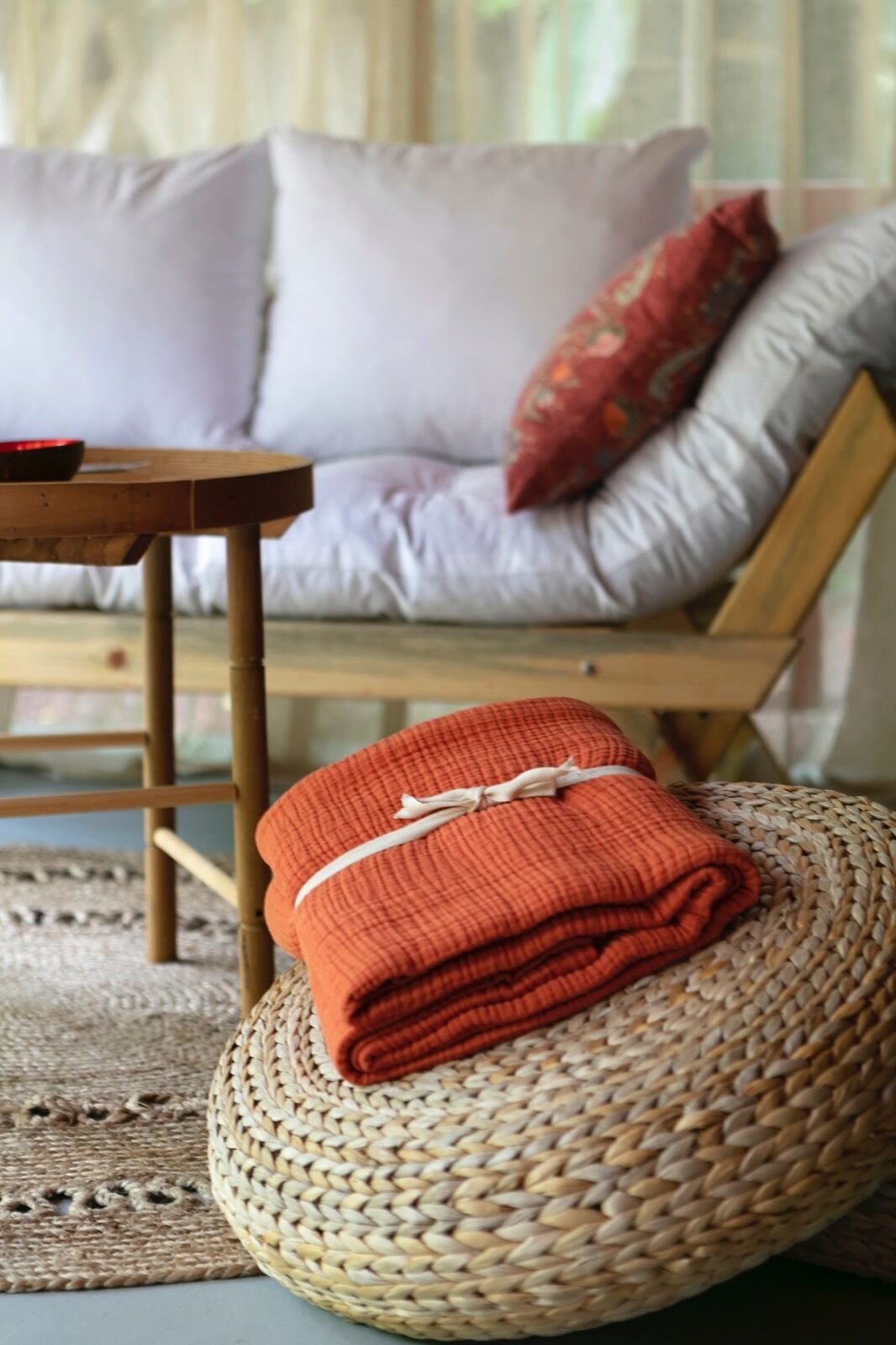 Un otomán de mimbre con una manta naranja doblada, una mesita de madera y un sofá con cojines estampados en una habitación.