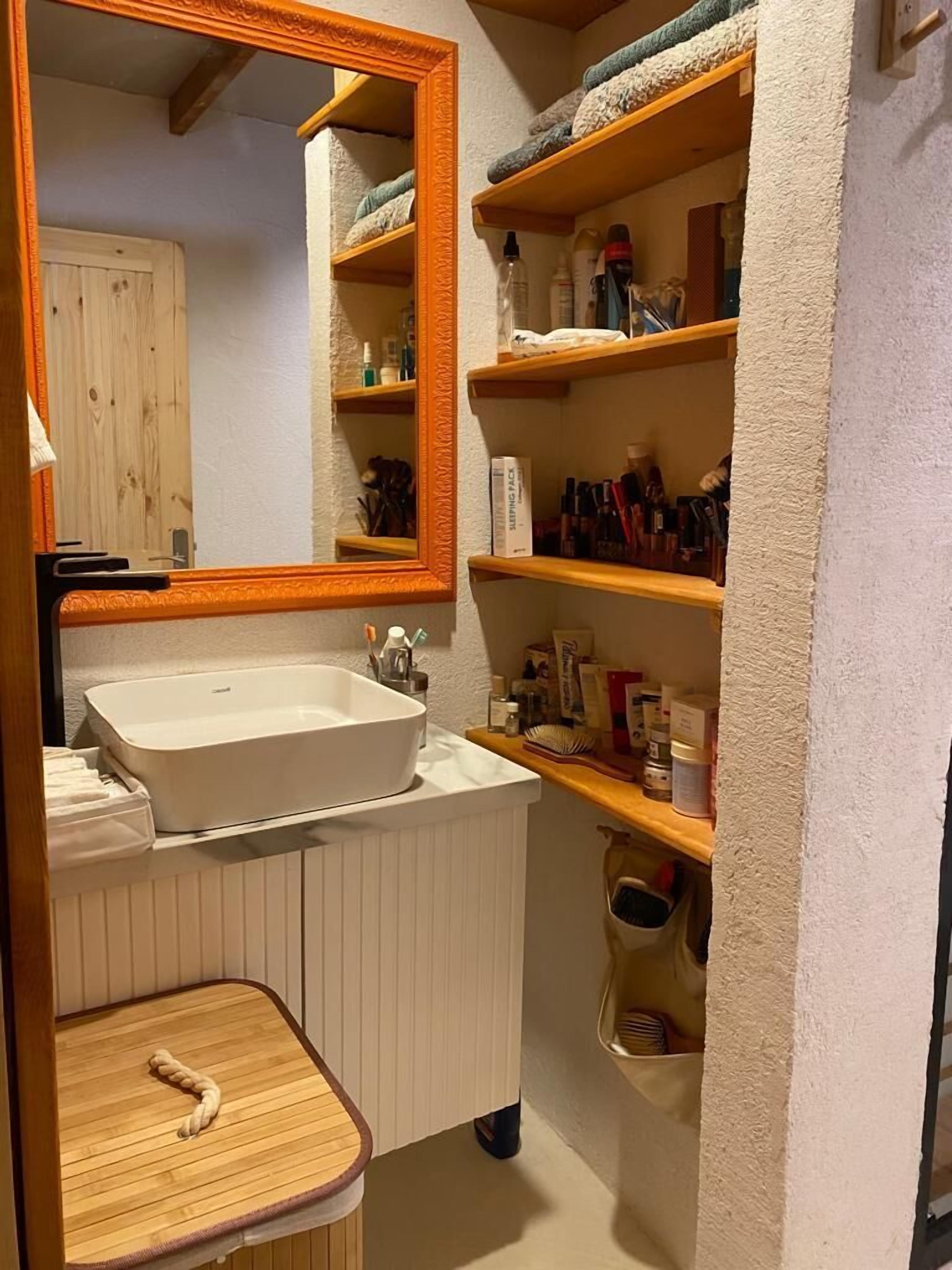 A bathroom with a white sink, a mirror, and shelves with toiletries.