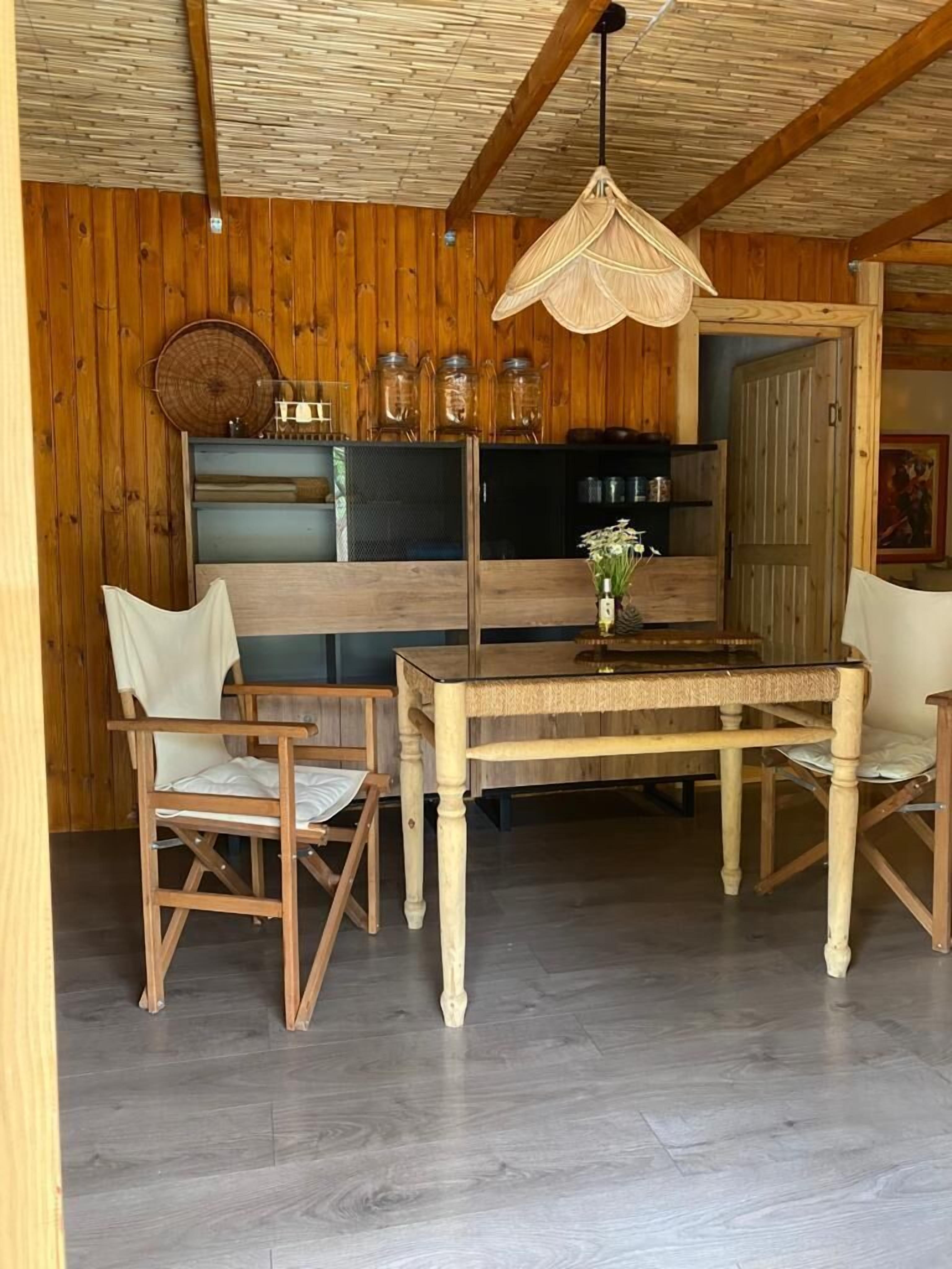 A wooden interior with a dining table, chairs, a hanging light fixture, and a kitchen area with jars and a refrigerator.