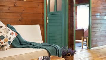 Interior acogedor de una cabaña de madera con piso de madera, una puerta verde, un sofá con cojines florales, una pequeña mesa de madera y un tapete tejido.