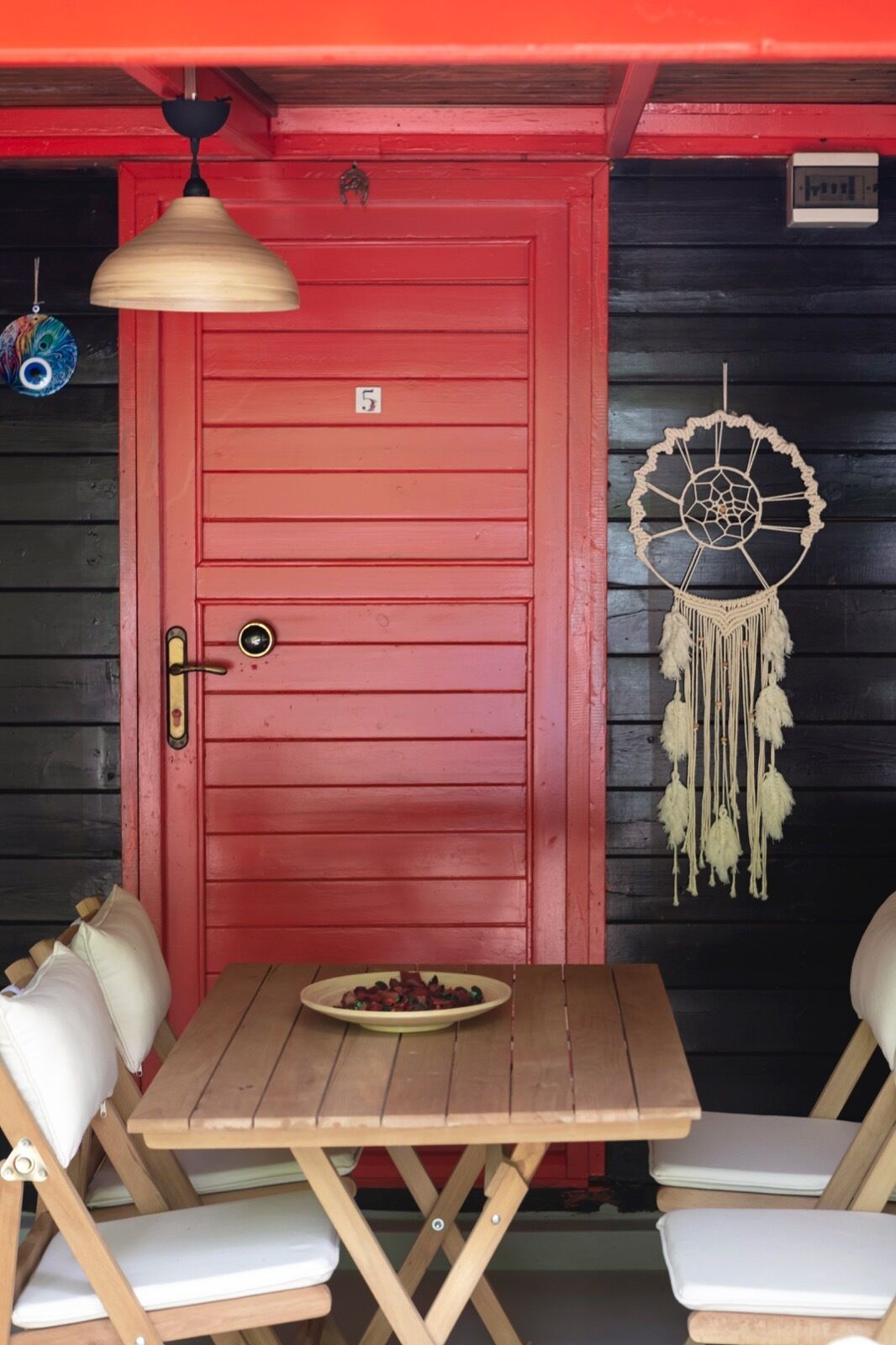 Una puerta roja con una placa de número, un atrapasueños y una mesa con sillas.
