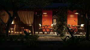 Un área de estar al aire libre con una mesa, sillas, un árbol pequeño y un edificio con puerta roja.