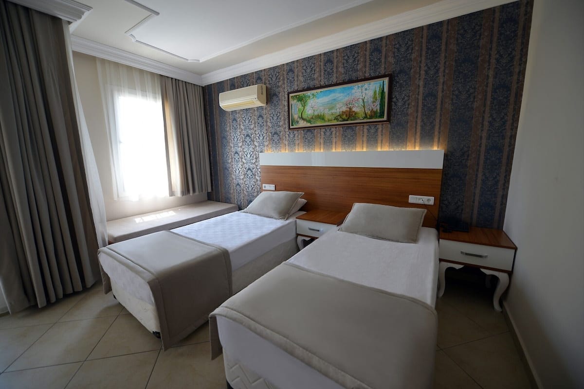 Quarto de hotel com duas camas de solteiro, uma janela grande com cortinas, um quadro na parede e cabeceira de madeira.