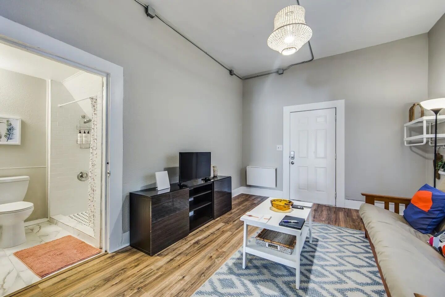 Una sala de estar moderna con piso de madera, un sofá blanco, una mesa de centro, un televisor sobre un mueble de madera oscura y una puerta que da a un baño.