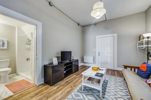 Ein modernes Wohnzimmer mit Holzboden, einem weißen Sofa, einem Couchtisch, einem Fernseher auf einem dunklen Holzschrank und einer Tür zu einem Badezimmer.