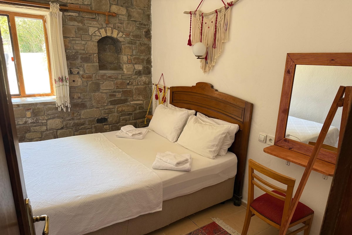 Un dormitorio con pared de piedra, una cama con ropa blanca, cabecera de madera, una pequeña silla de madera, un espejo y un tapete estampado.