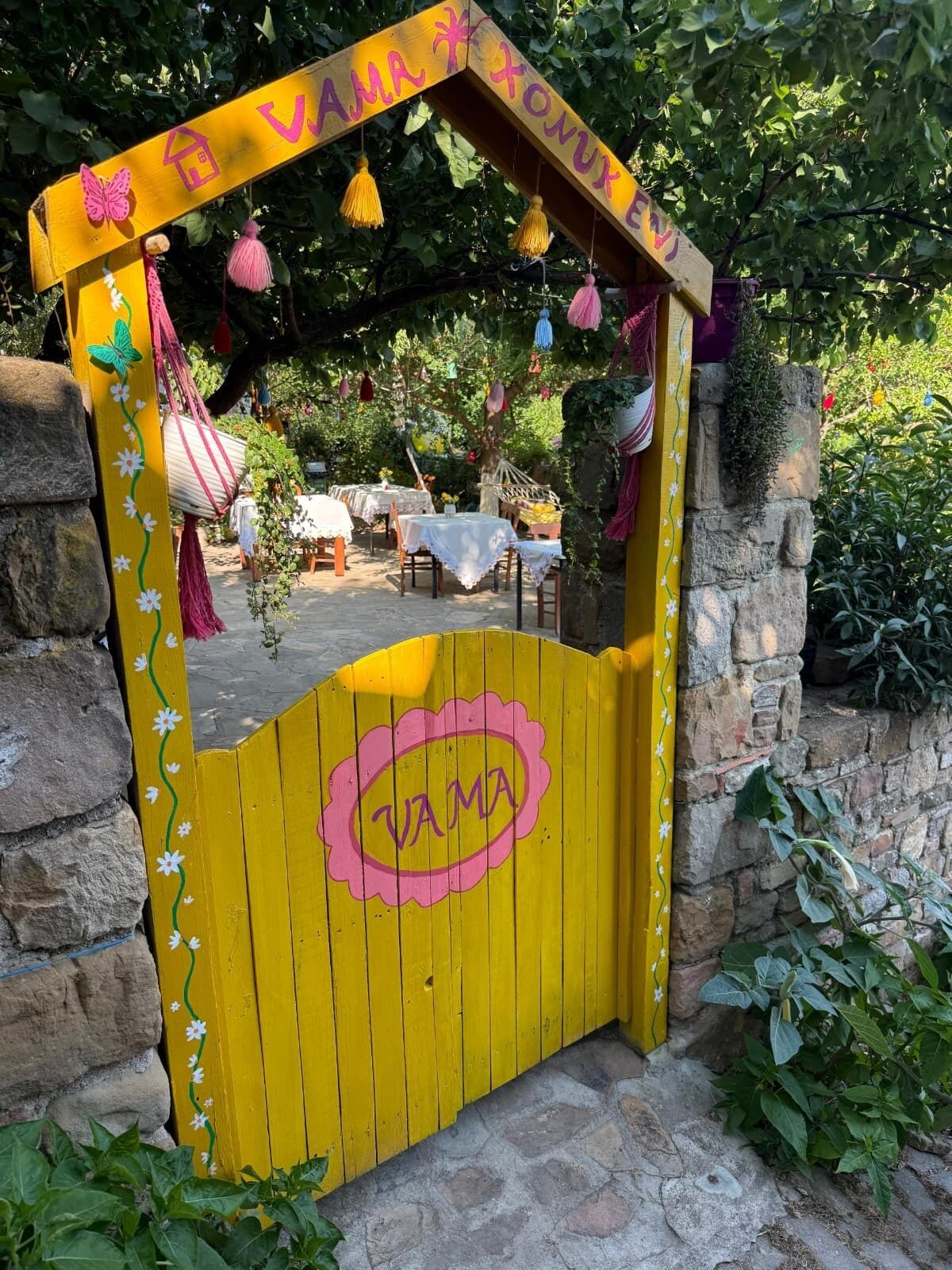 A yellow gate with 'VAMA' written on it, leading to an outdoor seating area with tables and chairs.