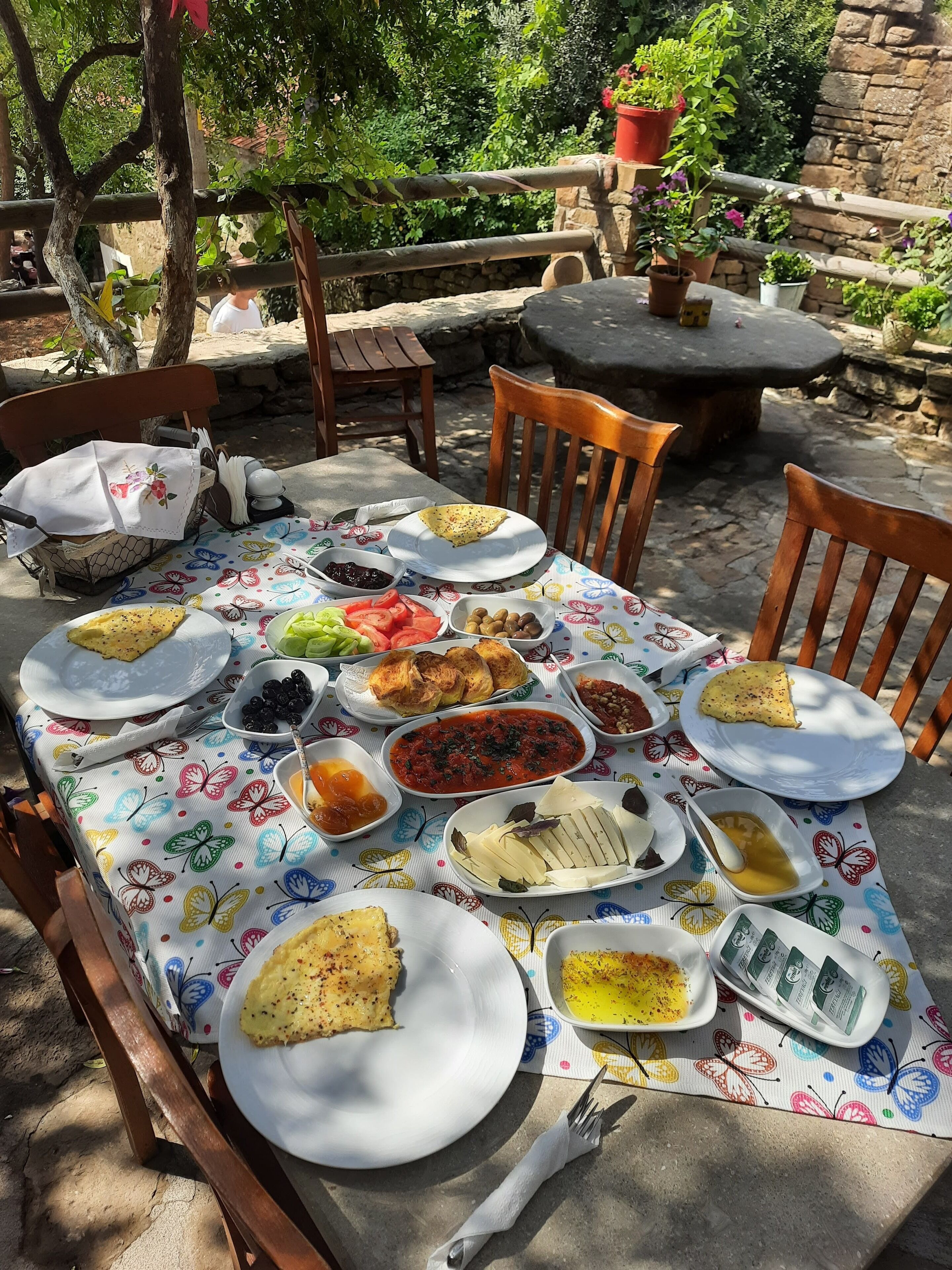 Un área de comedor al aire libre con diversos platos sobre una mesa, rodeada de sillas y un muro de piedra.