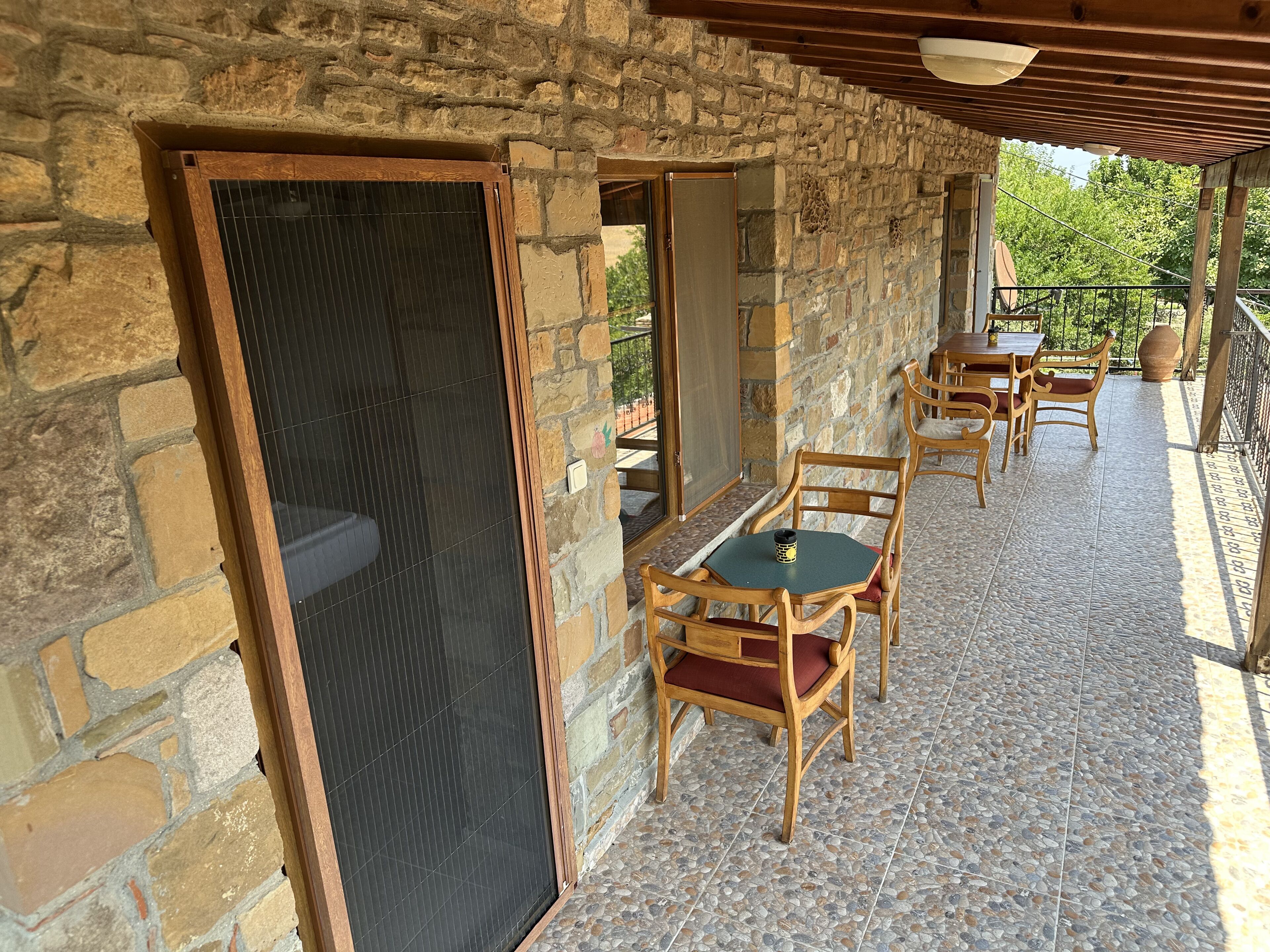 A stone-walled outdoor patio with wooden tables and chairs, a covered seating area, and a view of greenery.