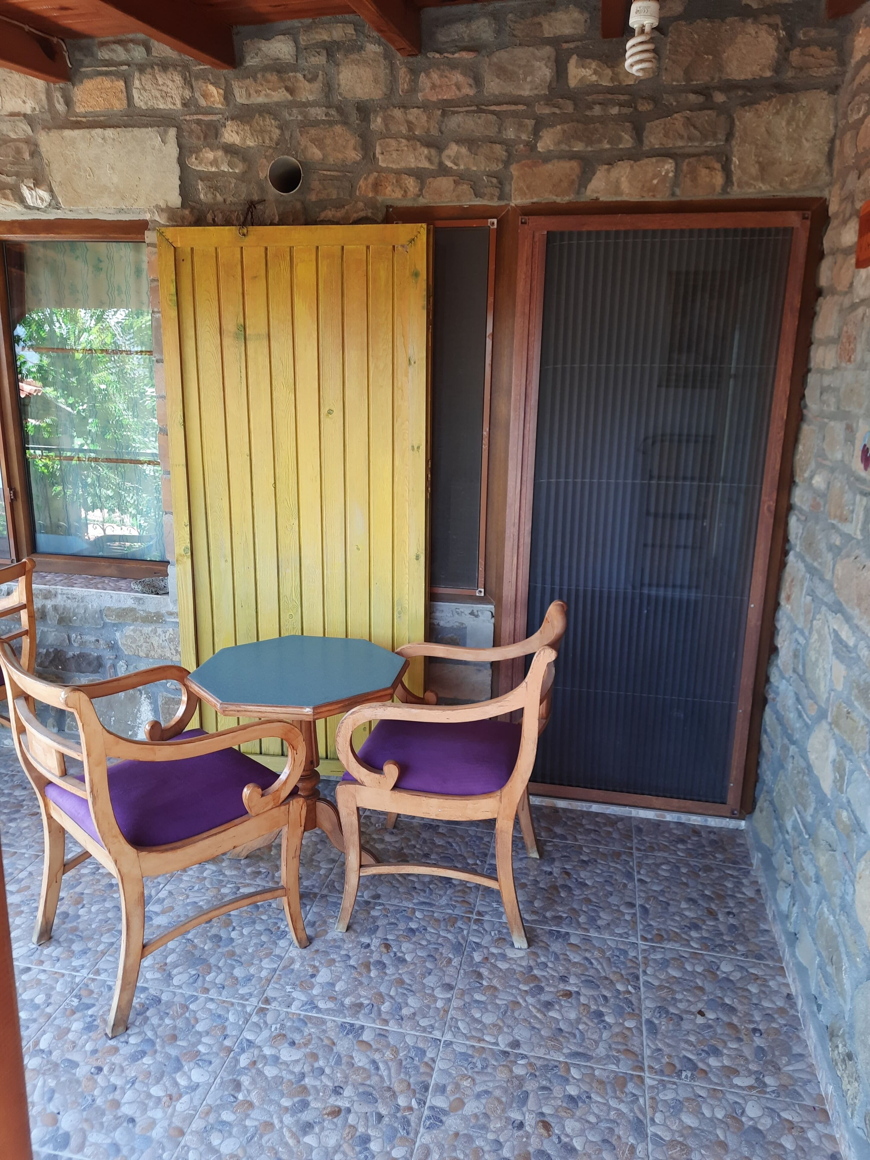 Un patio couvert avec un mur en pierre, une table en bois agrémentée de coussins violets et un paravent jaune.