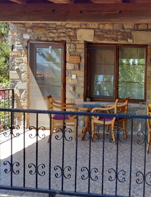 A stone-walled patio with a wooden table and chairs, a metal railing, and a view of greenery. - Vama Konuk Evi (Gökçeada)