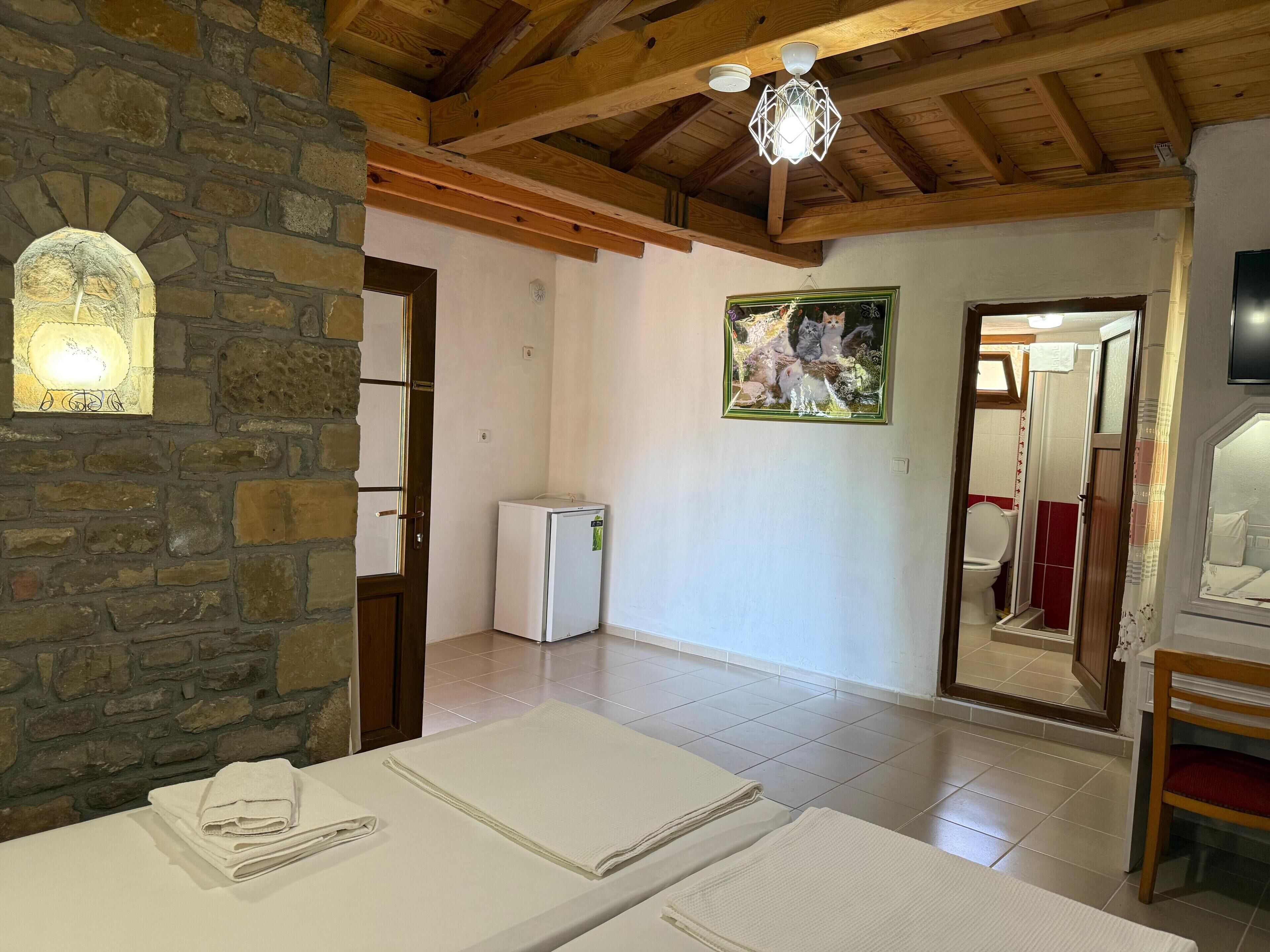 A room with a stone wall, wooden beams, a white bed with folded towels, a small white refrigerator, a painting, and a bathroom visible through an open door.