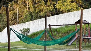 Una cadira de fil que connecta dues palanques en una zona del parc amb un parc infantil al fons.