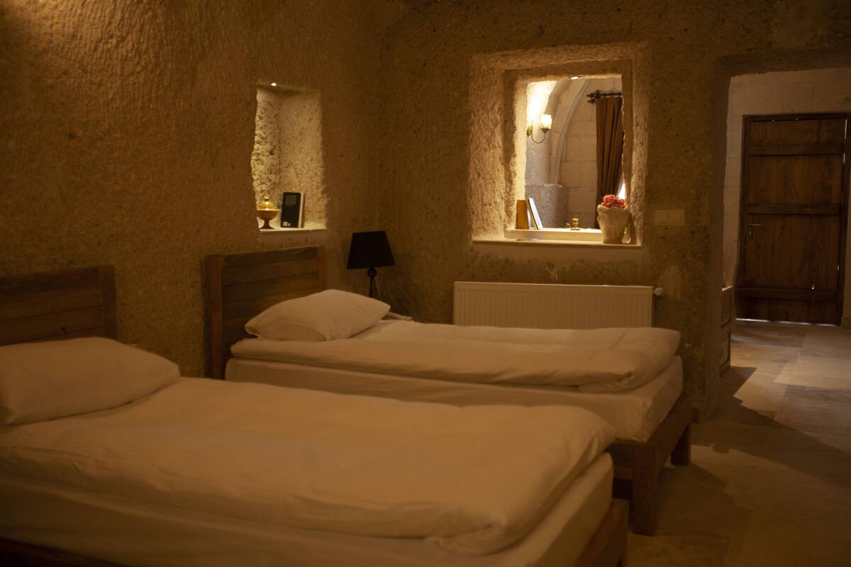 Quarto com duas camas de solteiro, janela com vista, radiador e porta de madeira.