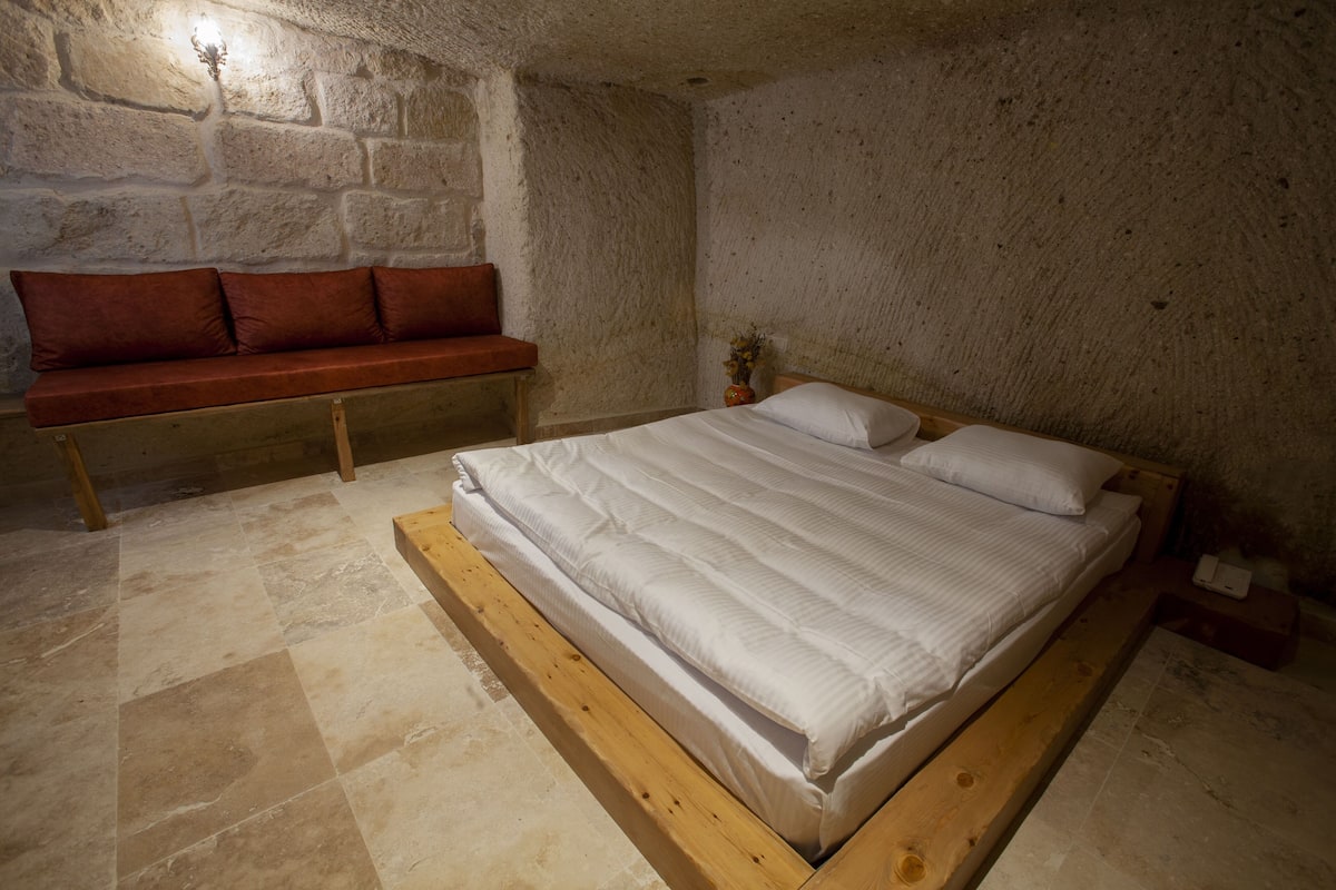 Quarto com estrutura de cama de madeira, um banquinho com almofadas e uma parede de pedra.