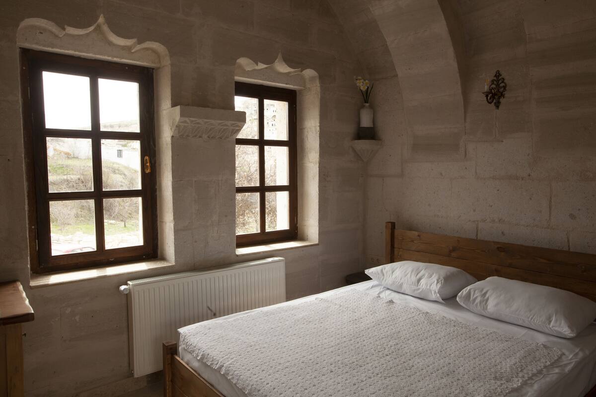 Quarto com cama de madeira, roupa de cama branca, duas janelas com cortinas e uma parede de pedra.