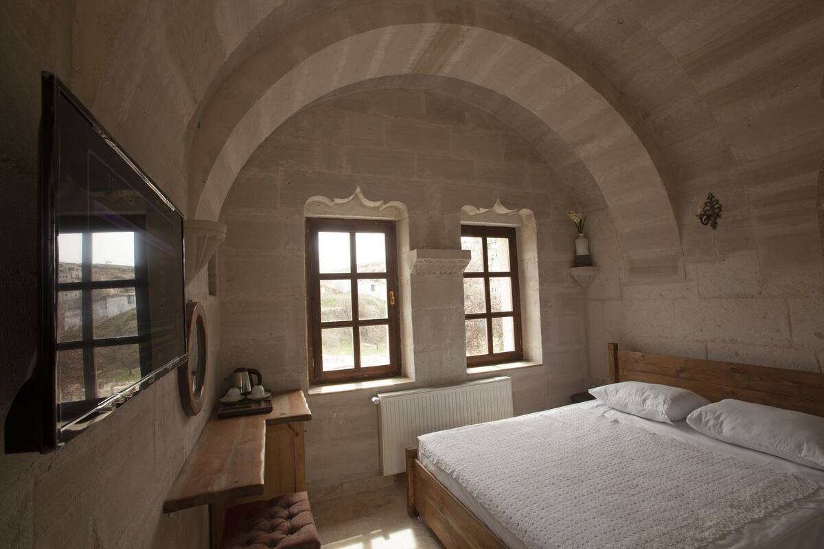 Quarto com cama de madeira, uma escrivaninha, duas janelas com cortinas e vista para o campo.