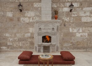 A stone fireplace with a fire, flanked by two red cushions and a small table with a teapot.
