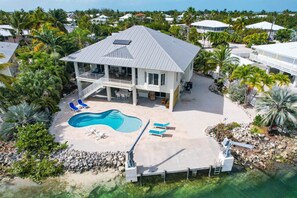 Une maison à deux étages avec une piscine, entourée de palmiers et offrant une vue dégagée sur l’océan.