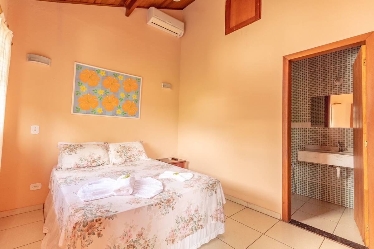 Une chambre à coucher comprenant un lit, une table de chevet, un climatiseur fixé au mur, une salle de bain avec un mur carrelé et une porte donnant accès à la salle de bain.