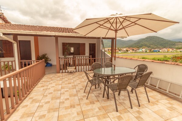 A covered patio with a round table and chairs, a large umbrella, and a view of a town and mountains in the background. - Pousada Luz do Sol (Paraty)