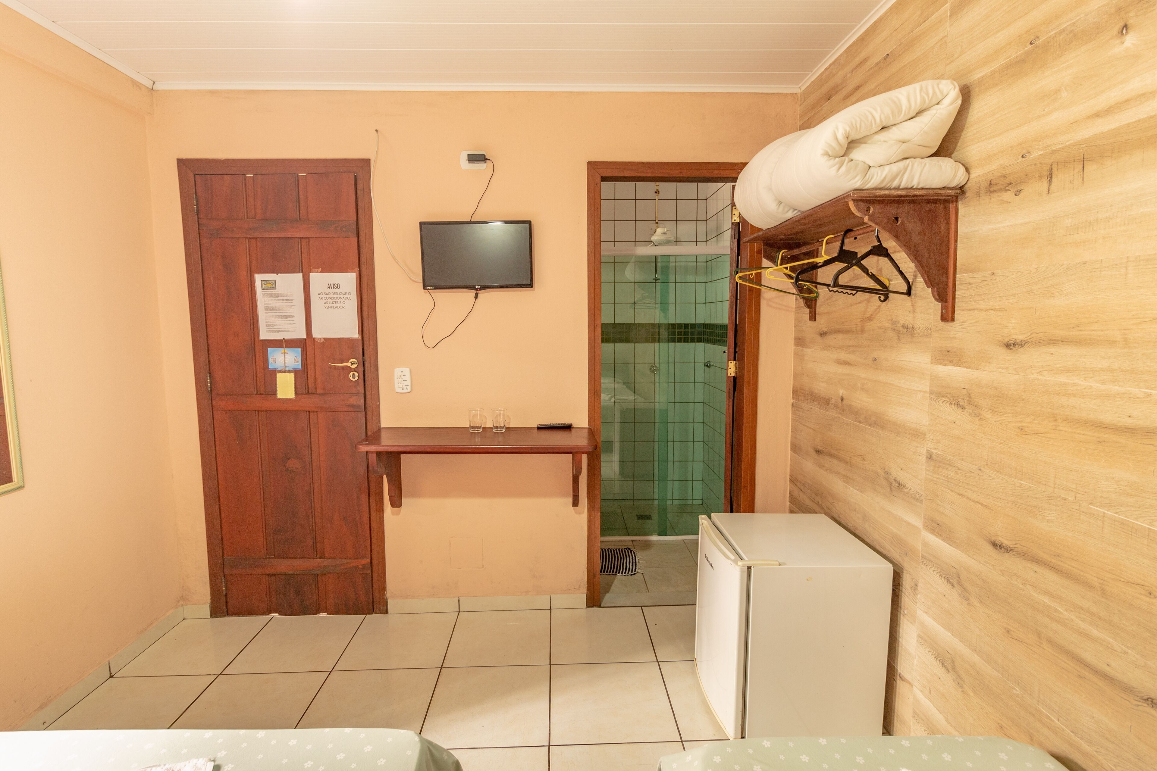 Een kleine kamer met een houten deur, een klein tafeltje met een televisie erboven, een witte koelkast en een handdoek aan een houten plank.