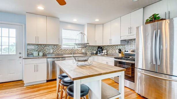 A modern kitchen with white cabinets, a stainless steel refrigerator, a wooden island with stools, and a window providing natural light.
