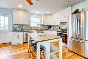 A modern kitchen with white cabinets, a stainless steel refrigerator, a wooden island with stools, and a window providing natural light. - Huge Beach House, Beautiful Sunsets | Sleeps 13! (Crystal Beach)