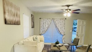 Ein Wohnzimmer mit einem weißen Sofa, einem Couchtisch und zwei Sesseln. Ein Deckenventilator und ein Fenster mit Vorhängen sind ebenfalls vorhanden.