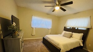 Ein Schlafzimmer mit Bett, Deckenventilator, Schminktisch und einem Fenster mit leichtem Vorhang.