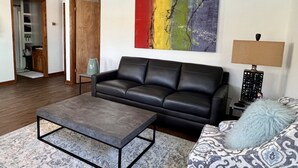 Un salón con un sofá de cuero negro, una mesita de centro gris, cojines estampados, un cuadro abstracto colorido y una lámpara.